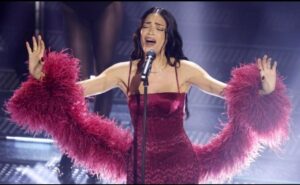 Elodie Di Patrizi on stage at Sanremo, singing in a red dress with pink, feathery sleeves. Sources and related content